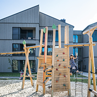 Wohngebäude mit Holzfassade. Vor dem Gebäude ein Spielplatz-Klettergerüst mit einem Kind