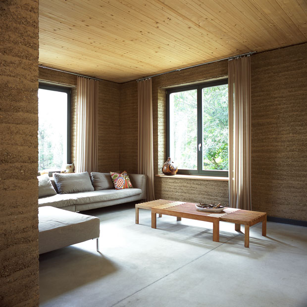 Wohnraum mit Sofa und zwei Fenstern. Die Decke ist aus hellem Holz, die Wände sind mit Lehm verputzt.