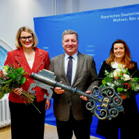 Gruppenbild mit drei Personen. Staatsminister Christian Bernreiter hält einen übergroßen Schlüssel