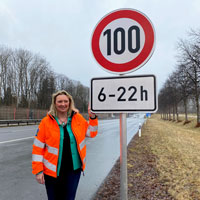 Verkehrsministerin Kerstin Schreyer steht mit oranger Warnweste neben einem Verkehrsschild an einer Autobahn. Das Verkehrsschild gibt ein Tempolimit von 100 km/h im Zeitraum von 6 bis 22 Uhr vor. 