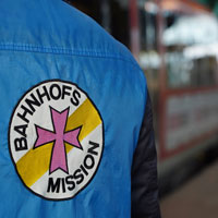 Eine Person mit einer blauen Jacke mit dem Logo der Bahnhofsmission steht an einem Bahnsteig. Im Hintergrund ist ein Zug zu sehen.
